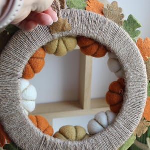 May include: A circular wreath wrapped in natural tan twine, decorated with small felt pumpkins in shades of orange, white, and olive green. The outer edge features felt oak leaves in orange, brown, and green, with a small twine loop for hanging.