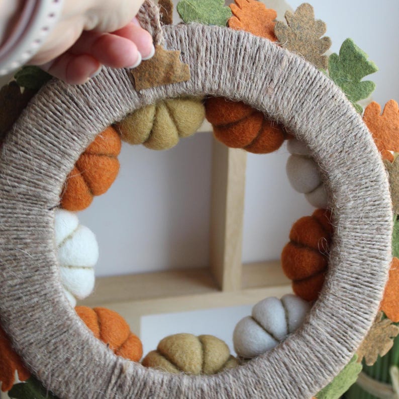 May include: A circular wreath wrapped in natural tan twine, decorated with small felt pumpkins in shades of orange, white, and olive green. The outer edge features felt oak leaves in orange, brown, and green, with a small twine loop for hanging.