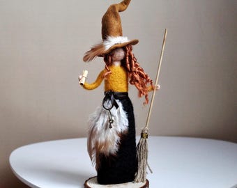 May include: A needle-felted doll featuring a tall brown pointed hat, a yellow long-sleeved top, and a black skirt adorned with white and brown feathers. The doll has long red hair, holds a small scroll and a broom, and stands on a wood slice.