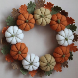 May include: A circular wreath constructed from felted wool elements, featuring a collection of small pumpkins in shades of orange, white, and muted tan, interspersed with numerous felt oak leaves in green, brown, and orange tones.