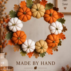 May include: A circular wreath made of felted pumpkins in shades of orange, white, and tan, interspersed with green and orange felt oak leaves. The wreath features a jute twine loop at the top for hanging. Text below reads MADE BY HAND ONE PIECE AT A TIME.