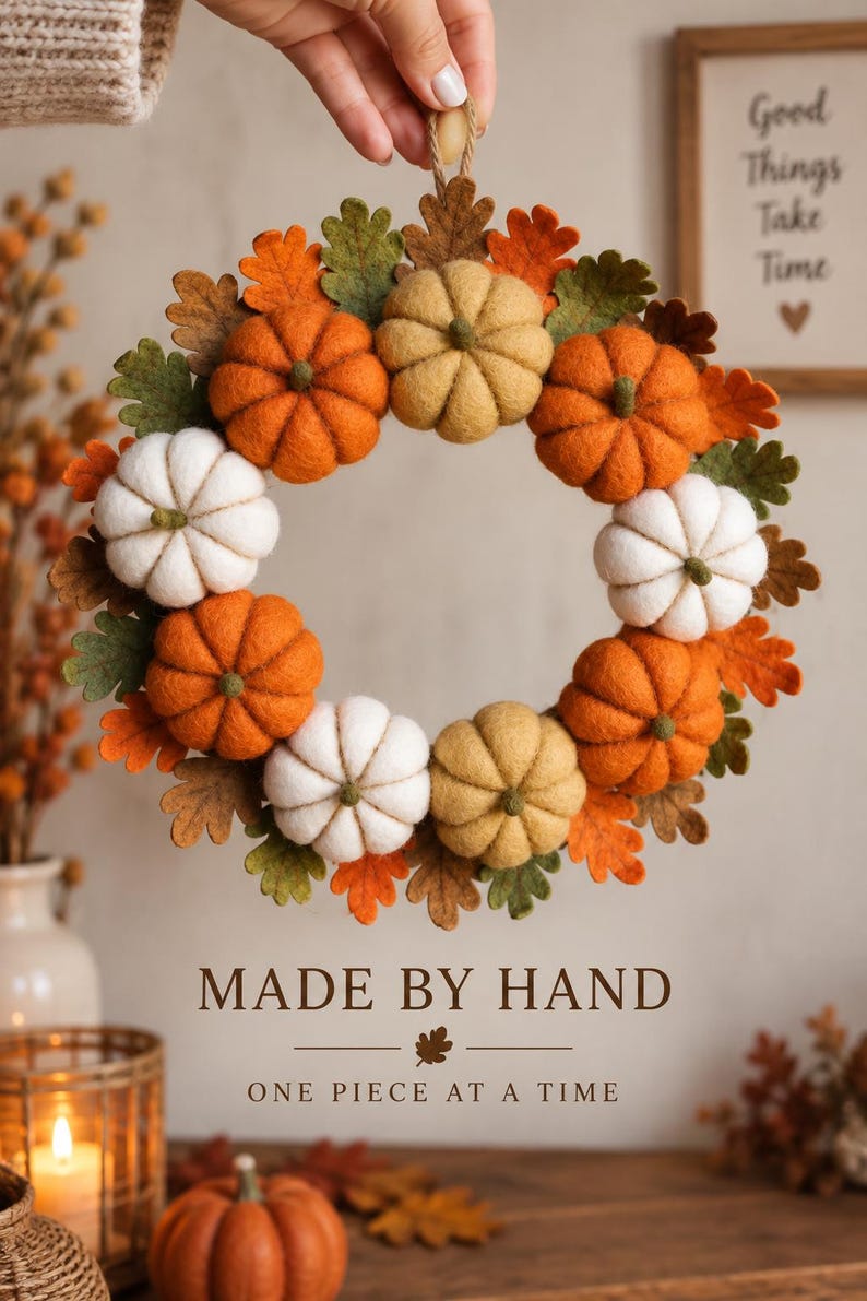 May include: A circular wreath made of felted pumpkins in shades of orange, white, and tan, interspersed with green and orange felt oak leaves. The wreath features a jute twine loop at the top for hanging. Text below reads MADE BY HAND ONE PIECE AT A TIME.