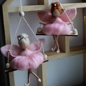 May include: Two handmade fairy ornaments. Each fairy has pink wings and a pink dress with glitter. They are suspended from a wooden swing with white string and wooden beads. The fairies have brown hair and are decorated with pearls.