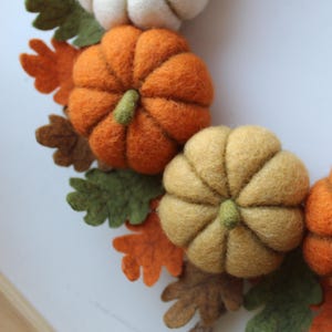 May include: A decorative wreath featuring felted wool pumpkins in shades of orange, yellow, and white, interspersed with felted leaves in green, brown, and orange. The pumpkins have small green stems and a textured, soft appearance.