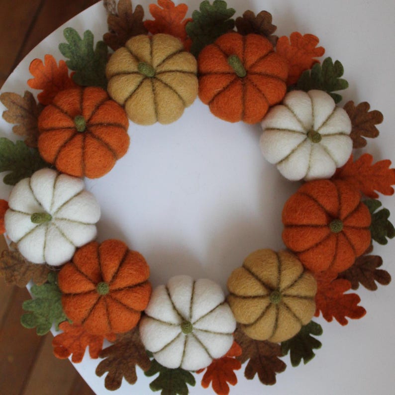 May include: A circular wreath made of felted wool pumpkins in shades of orange, white, and mustard yellow, interspersed with felt oak leaves in green, brown, and orange. The pumpkins are arranged in a ring, each featuring a small green stem.