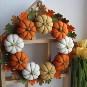 May include: A circular wreath featuring felted pumpkins in shades of orange, white, and muted yellow, interspersed with felt oak leaves in green, brown, and orange. The wreath is mounted on a light-coloured wooden frame shaped like a house.