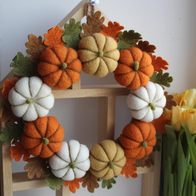 May include: A circular wreath featuring felted pumpkins in shades of orange, white, and muted yellow, interspersed with felt oak leaves in green, brown, and orange. The wreath is mounted on a light-coloured wooden frame shaped like a house.