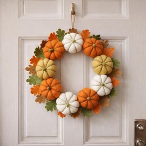 May include: A circular wreath constructed from felted wool pumpkins in shades of orange, yellow, and white, interspersed with green and brown felted oak leaves. The decorative piece is suspended by a thin twine loop from a metal hook on a door.