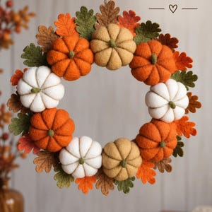 May include: A circular wreath featuring felt pumpkins in shades of orange, white, and tan, interspersed with felt oak leaves in green, brown, and orange. A jute twine loop is attached at the top with text that reads READY TO HANG with a heart.