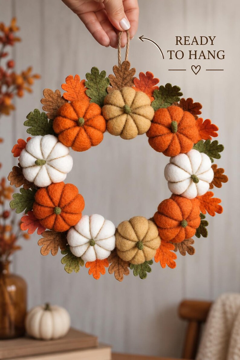 May include: A circular wreath featuring felt pumpkins in shades of orange, white, and tan, interspersed with felt oak leaves in green, brown, and orange. A jute twine loop is attached at the top with text that reads READY TO HANG with a heart.