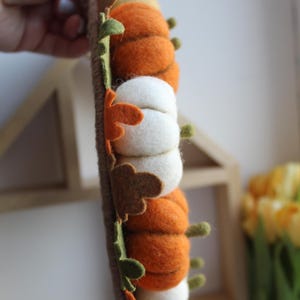 May include: A vertical garland featuring alternating felt pumpkins in shades of orange and white, accented with small felt leaves in green, brown, and orange. The pumpkins are attached to a brown twine-wrapped base, creating a textured decoration.