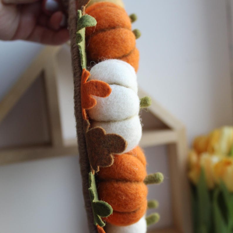May include: A vertical garland featuring alternating felt pumpkins in shades of orange and white, accented with small felt leaves in green, brown, and orange. The pumpkins are attached to a brown twine-wrapped base, creating a textured decoration.