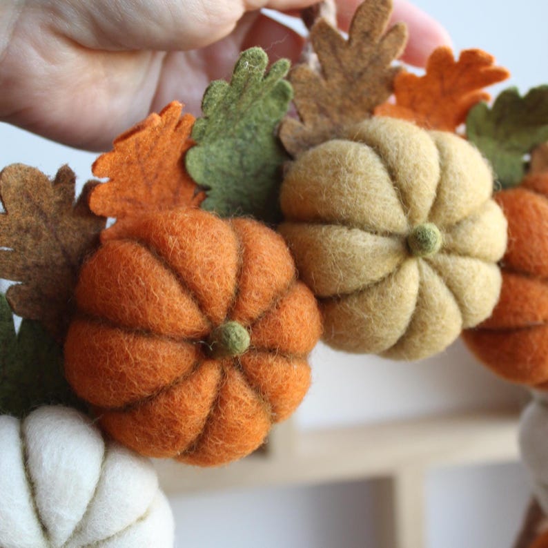 May include: A decorative wreath featuring needle-felted pumpkins in shades of orange, cream, and muted yellow, interspersed with felted autumn leaves in green, brown, and orange tones. The pumpkins have distinct ribbed textures and small stems.