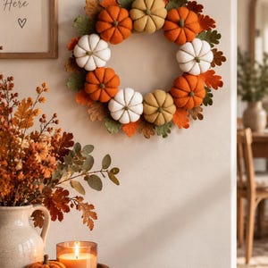 May include: A circular wreath featuring felt pumpkins in shades of orange, white, and muted yellow, interspersed with green and orange felt leaves. The wreath is suspended by a jute cord and hangs on a wall above a wooden table with decor.