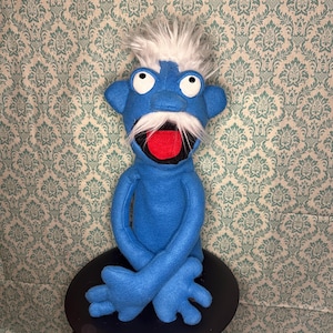 May include: A blue felt puppet with a white mustache and hair, a red mouth, and large white eyes. The puppet is sitting on a black stool in front of a patterned wallpaper.