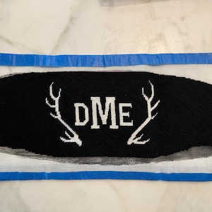 May include: A rectangular needlepoint canvas featuring a black background with white stitched letters DME in the centre, flanked by white deer antler silhouettes. The canvas is bordered by blue tape on the top and bottom edges.