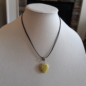 May include: A heart-shaped pendant necklace. The pendant is a light green color with carved lines. The necklace has a black cord and a silver-colored metal clasp. The necklace is displayed on a white mannequin.