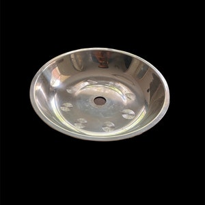 May include: A shiny, silver metal bowl with a circular shape and a central hole. The bowl has a series of decorative indentations around the base. The bowl is set against a black background.