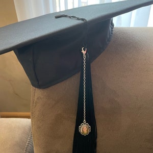 May include: Black graduation cap with a black tassel and a silver charm with a photo of a person. The charm is attached to the tassel with a silver chain.