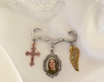 May include: A silver brooch with a delicate, branch-like design. It features three charms: a gold angel wing, a copper cross, and a silver oval frame with a photo of a person inside.