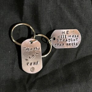 May include: Two silver keychains with inspirational messages. One keychain says "Take the high road" with a peace sign. The other keychain says "HE will make your paths straight" with a small cross.
