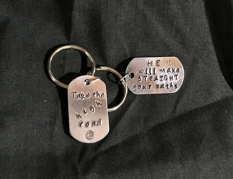 May include: Two silver keychains with inspirational messages. One keychain says "Take the high road" with a peace sign. The other keychain says "HE will make your paths straight" with a small cross.