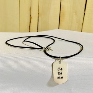 May include: A silver dog tag necklace with a black leather cord. The dog tag has the words "Ja Ro me" engraved on it.