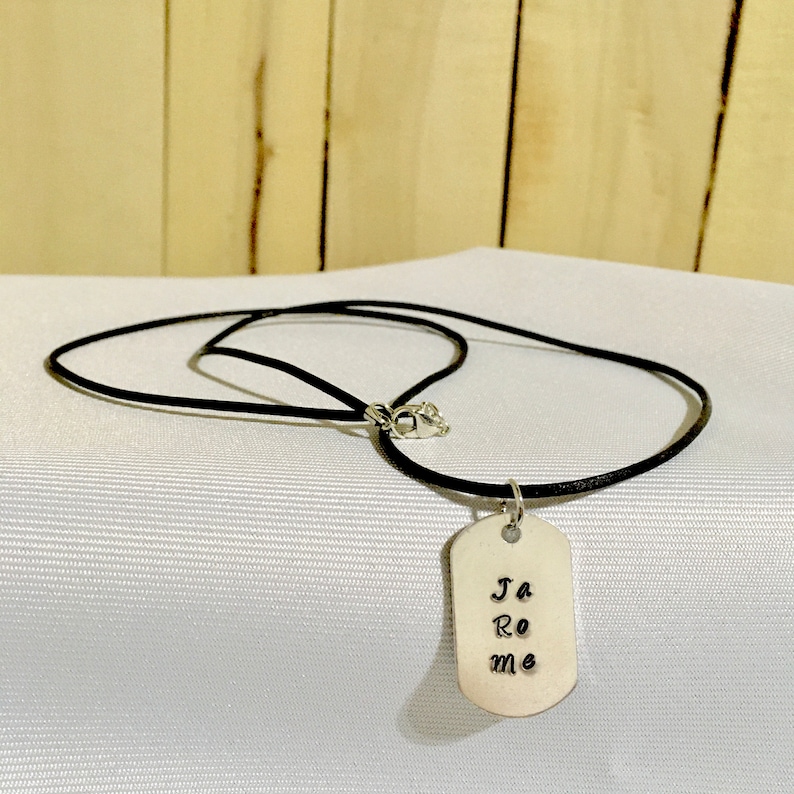 May include: A silver dog tag necklace with a black leather cord. The dog tag has the words "Ja Ro me" engraved on it.