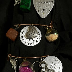 May include: Three silver charm bracelets with various charms, including a heart charm with the text "I love you to the moon & back", a guitar pick charm with the text "Life has changed but not ended", a square charm with brown glitter, a glass orb charm with pink glitter, a dolphin charm, and a heart charm with the text "I can do this".