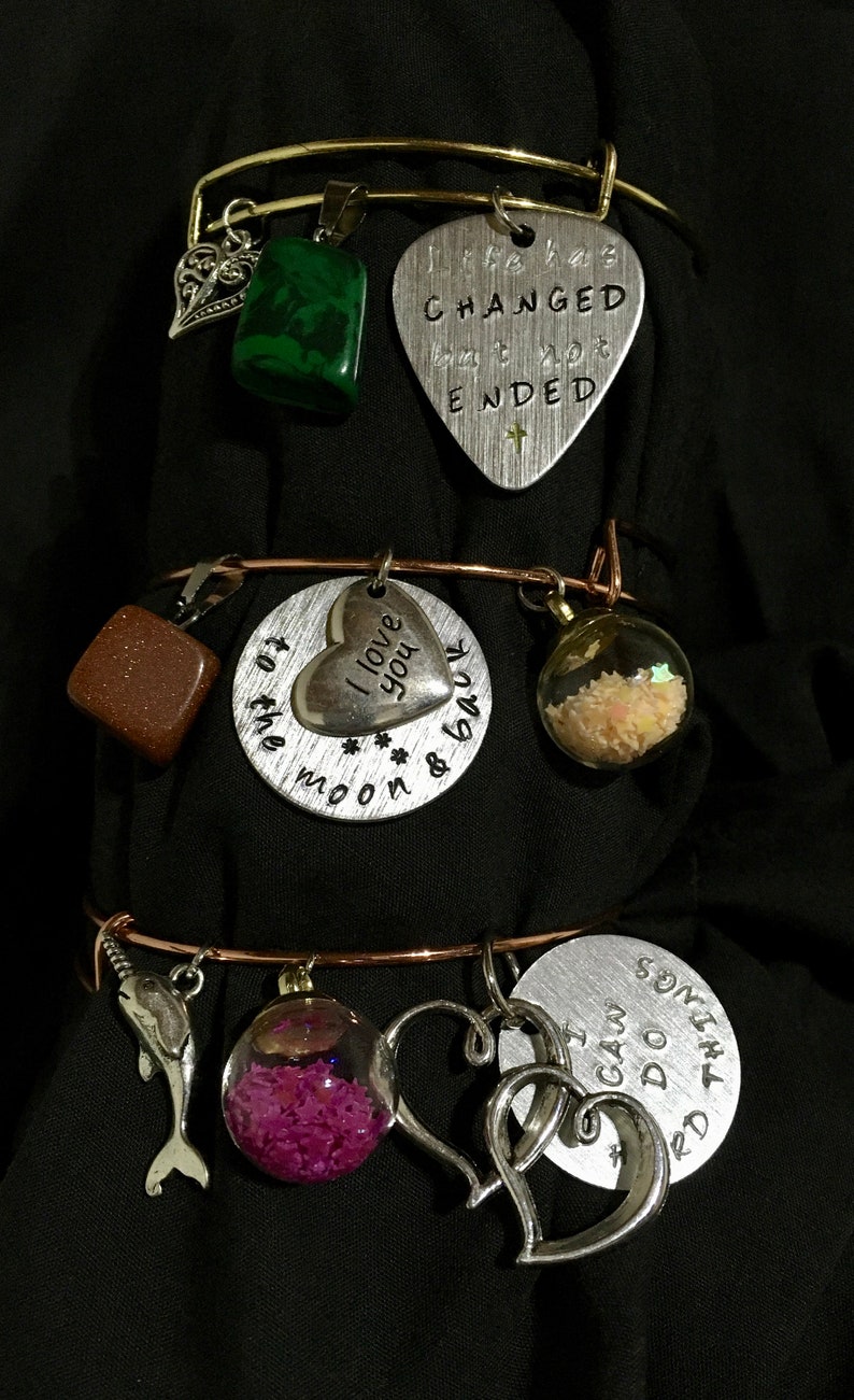 May include: Three silver charm bracelets with various charms, including a heart charm with the text "I love you to the moon & back", a guitar pick charm with the text "Life has changed but not ended", a square charm with brown glitter, a glass orb charm with pink glitter, a dolphin charm, and a heart charm with the text "I can do this".