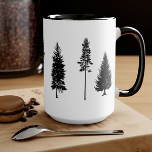 May include: White ceramic mug with a black rim and handle. The mug features a black silhouette design of three pine trees.