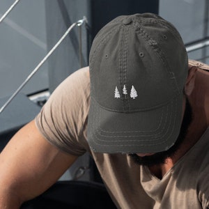 May include: A dark gray distressed baseball cap with a white embroidered design of three trees. The cap is worn by a person sitting on a set of stairs.