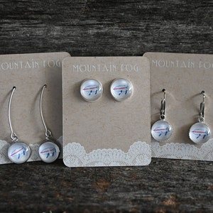 May include: Three pairs of earrings with a blue and red buffalo logo on the front. The earrings are displayed on brown cards with the text "Mountain Fog" printed on them.