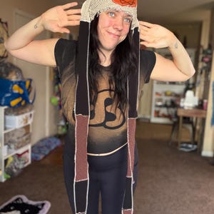 May include: A long, hooded headpiece featuring a beige crocheted cap with a circular orange patch displaying a dark spiral design. Two long, rectangular fabric tails hang down, constructed from alternating black and brown panels with visible white stitching.