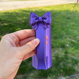May include: A purple decorative bow-shaped object. The bow is attached to a rectangular base. The object is held in a hand against a background of green grass and a residential neighborhood.