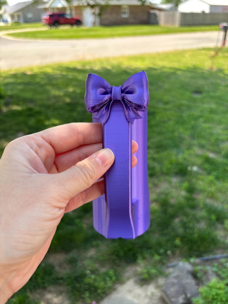 May include: A purple decorative bow-shaped object. The bow is attached to a rectangular base. The object is held in a hand against a background of green grass and a residential neighborhood.