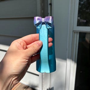 May include: A 3D-printed teal and purple bow-shaped bottle stopper. The bow is a gradient of purple to teal, and the stopper has a rectangular handle. The object is held in front of a white and gray background.