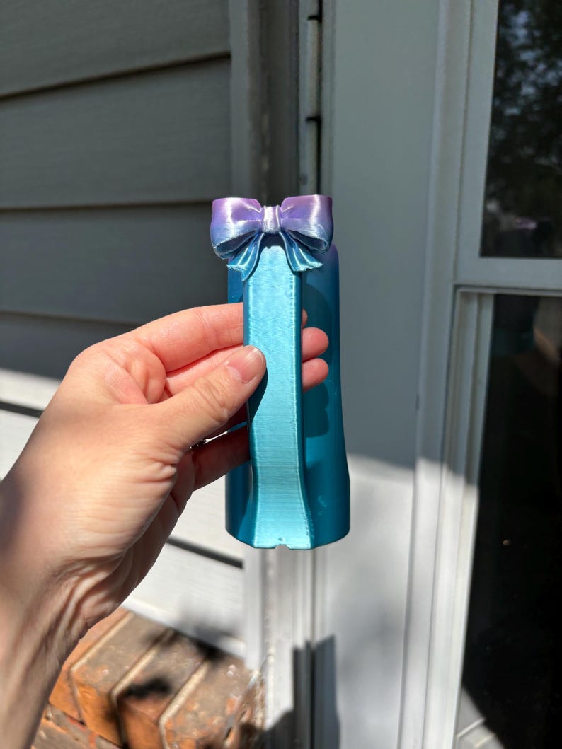 May include: A 3D-printed teal and purple bow-shaped bottle stopper. The bow is a gradient of purple to teal, and the stopper has a rectangular handle. The object is held in front of a white and gray background.