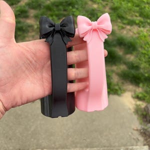 May include: Two decorative bow-shaped objects, one black and one pink, are held up. The bows are at the top of a rectangular column. The pink bow is a lighter shade than the black one. The background is blurred green grass.