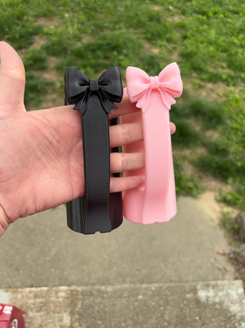 May include: Two decorative bow-shaped objects, one black and one pink, are held up. The bows are at the top of a rectangular column. The pink bow is a lighter shade than the black one. The background is blurred green grass.