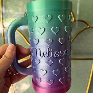 K&ouml;nnte beinhalten: Ein personalisierter Becher mit einem Farbverlauf von Gr&uuml;n &uuml;ber Lila bis hin zu Rosa. Der Becher ist mit herzf&ouml;rmigen Designs und dem Namen "Melissa" in Schreibschrift verziert.