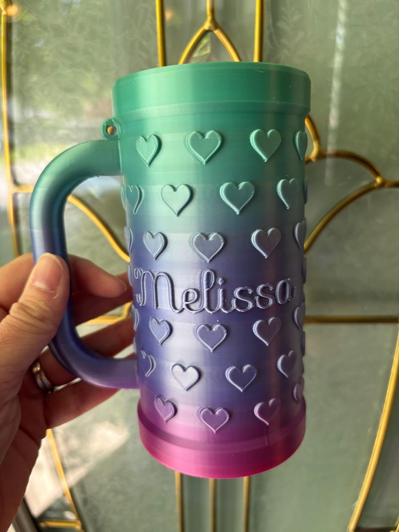 K&ouml;nnte beinhalten: Ein personalisierter Becher mit einem Farbverlauf von Gr&uuml;n &uuml;ber Lila bis hin zu Rosa. Der Becher ist mit herzf&ouml;rmigen Designs und dem Namen "Melissa" in Schreibschrift verziert.