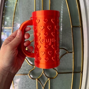 K&ouml;nnte beinhalten: Eine leuchtend orangefarbene Tasse mit Henkel, bedeckt mit herzf&ouml;rmigen Aussparungen. Der Name "Kenya" ist auf der Seite aufgedruckt. Die Tasse wird vor einer Glast&uuml;r gehalten.