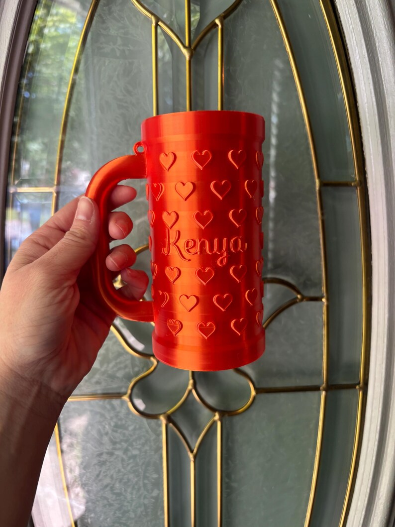 K&ouml;nnte beinhalten: Eine leuchtend orangefarbene Tasse mit Henkel, bedeckt mit herzf&ouml;rmigen Aussparungen. Der Name "Kenya" ist auf der Seite aufgedruckt. Die Tasse wird vor einer Glast&uuml;r gehalten.