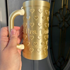 K&ouml;nnte beinhalten: Ein goldfarbener Becher mit Henkel, verziert mit Herzformen. Der Becher tr&auml;gt die Aufschrift "Mrs. Vail" auf der Vorderseite. Der Becher wird von einer Person gehalten.