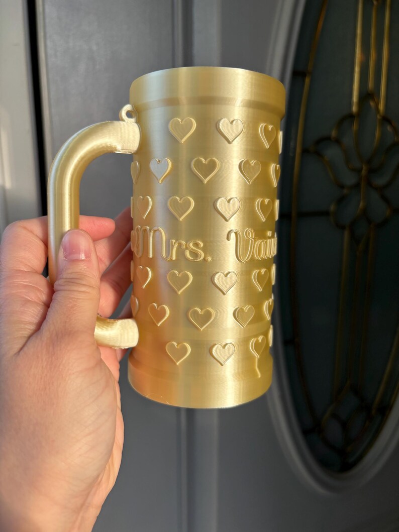 K&ouml;nnte beinhalten: Ein goldfarbener Becher mit Henkel, verziert mit Herzformen. Der Becher tr&auml;gt die Aufschrift "Mrs. Vail" auf der Vorderseite. Der Becher wird von einer Person gehalten.