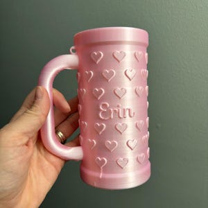 K&ouml;nnte beinhalten: Ein rosa Becher mit Henkel und zylindrischem K&ouml;rper. Der Becher ist mit gepr&auml;gten Herzen und dem Namen "Erin" verziert. Der Becher besteht aus einem 3D-gedruckten Material.