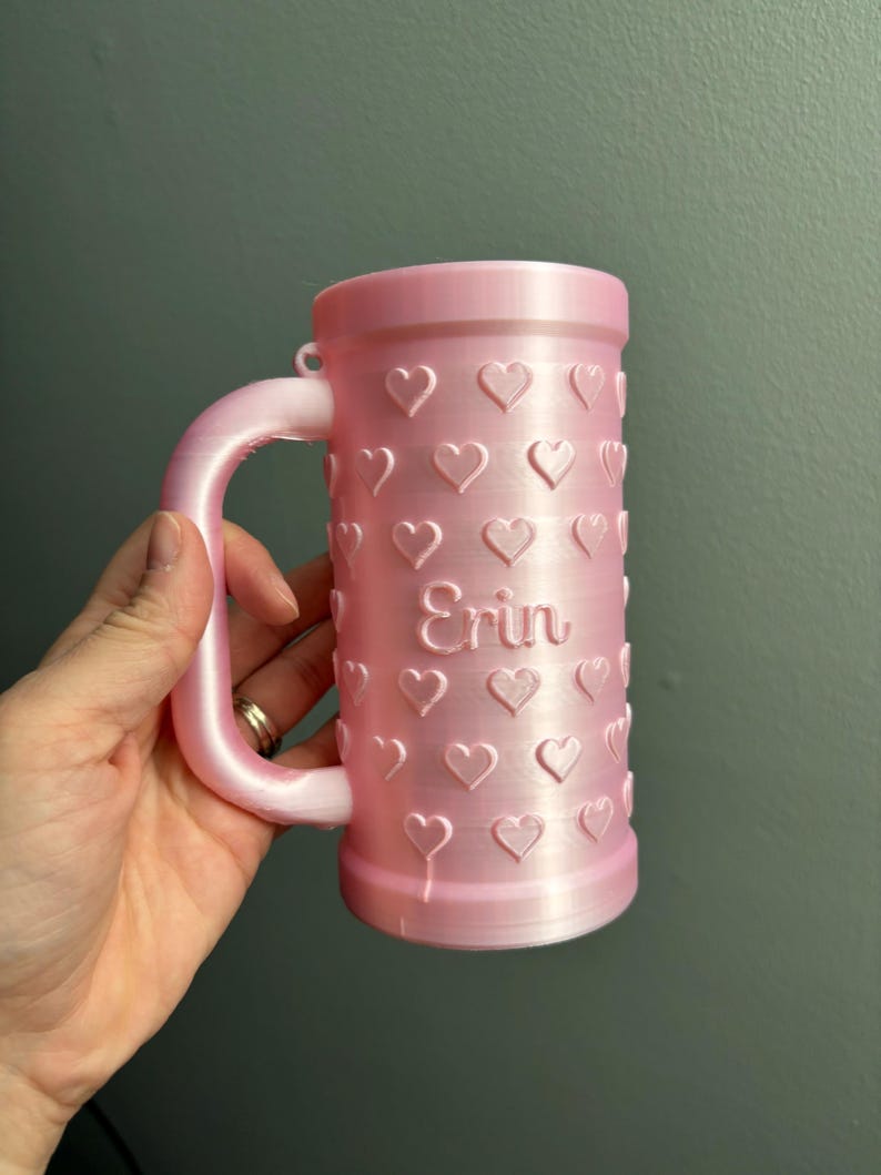 K&ouml;nnte beinhalten: Ein rosa Becher mit Henkel und zylindrischem K&ouml;rper. Der Becher ist mit gepr&auml;gten Herzen und dem Namen "Erin" verziert. Der Becher besteht aus einem 3D-gedruckten Material.