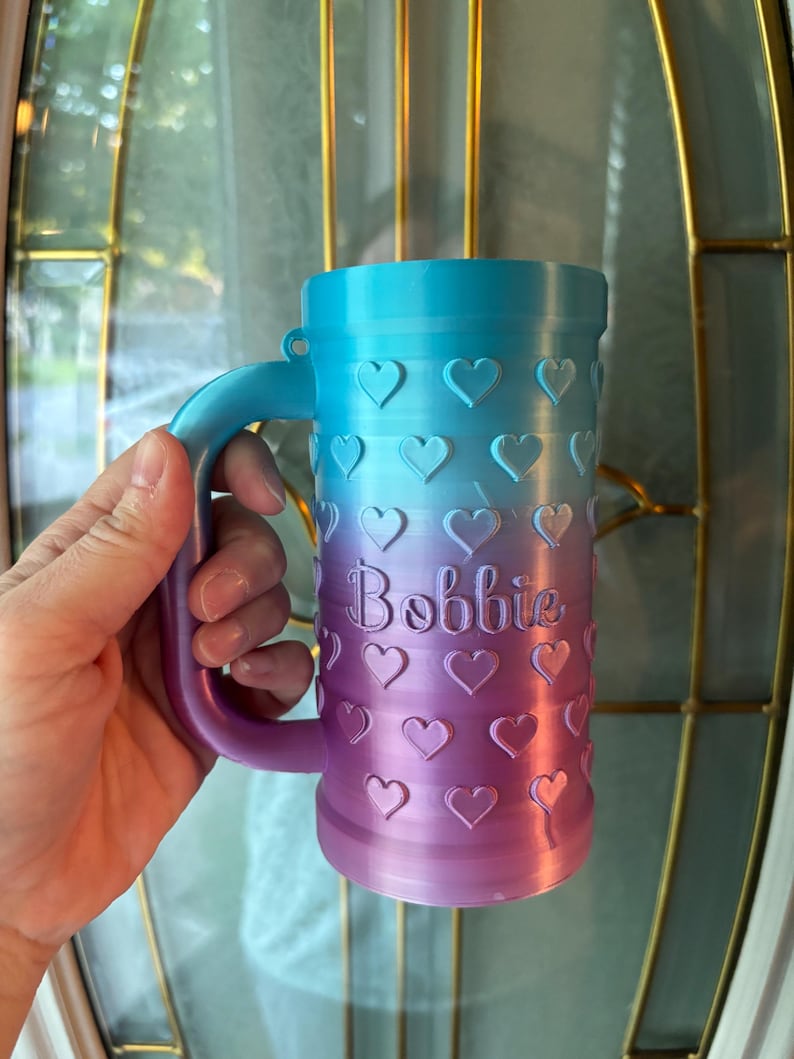 K&ouml;nnte beinhalten: Ein blau-lila Farbverlaufskrug mit Henkel, verziert mit gepr&auml;gten Herzen und dem Namen "Bobbie". Der Krug hat eine zylindrische Form und einen gro&szlig;en Henkel. Der Krug wird vor einem Fenster gehalten.