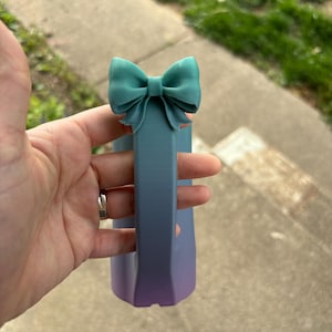 May include: A teal bow-shaped accent tops a gradient-colored, rectangular object. The object transitions from blue to purple. The bow is a solid color, while the object has a smooth, matte finish. The object's shape is geometric.